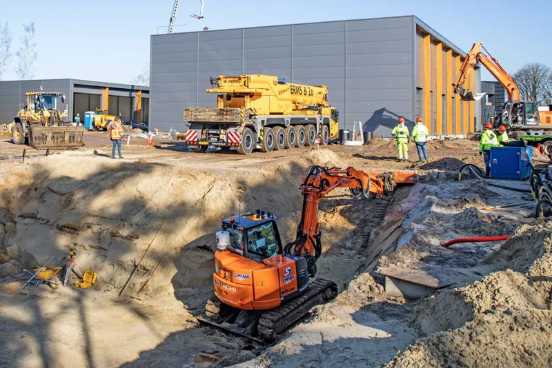 Graafmachines en medewerkers bij grondwerk naast een bedrijfshal.