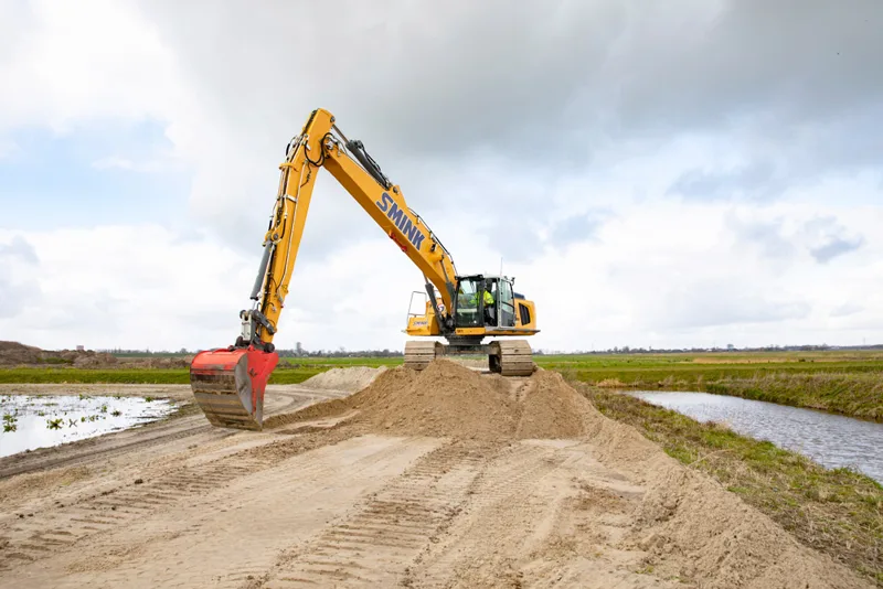 Graafmachine verplaatst zand tussen water en grasland.