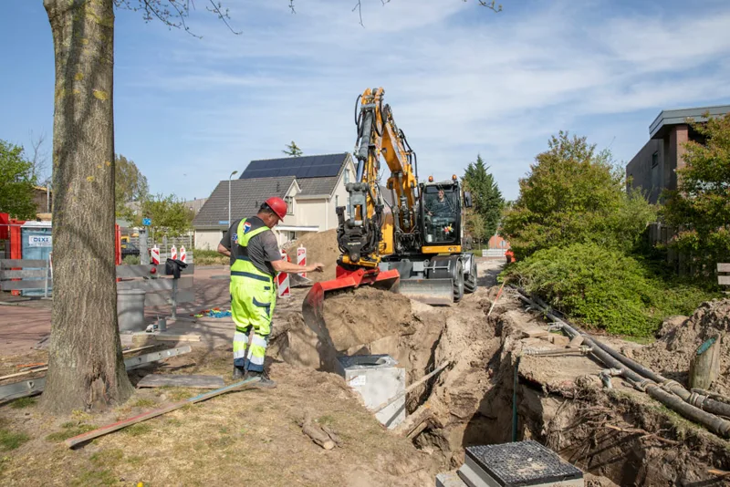 Medewerker en graafmachine bij een open sleuf in een woonwijk.