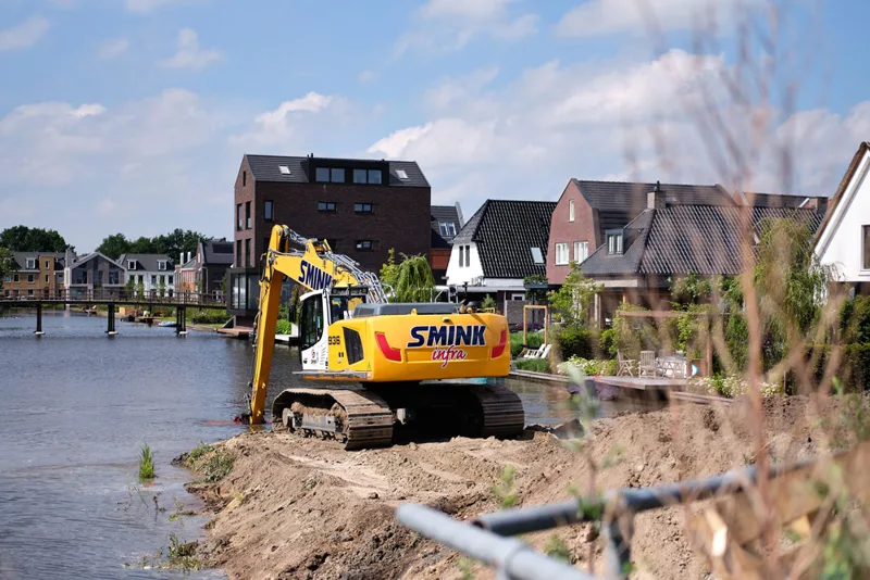 Graafmachine werkt aan de oever van een watergang naast woningen.